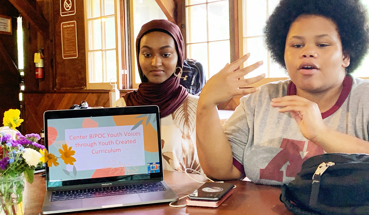 Two youth talk with a laptop on the table whose screen read Center BIPOC youth voices through youth created curriculum